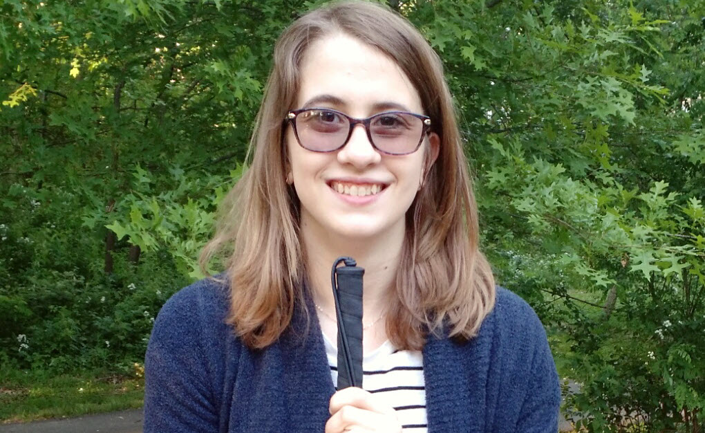 A smiling young woman with brown hair standing in a garden. She is wearing a blue sweater, dark-rimmed glasses , and is holding a white cane.