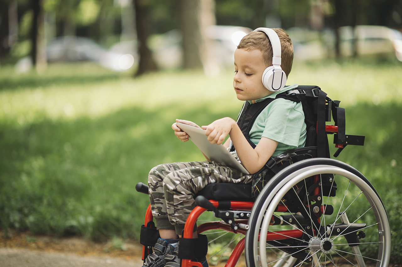 Young student in wheelchair using headphones and device outdoors
