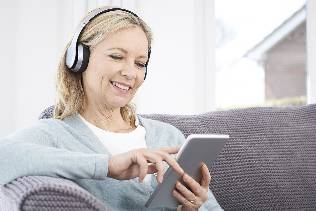 Senior woman using tablet and headphones on home sofa.