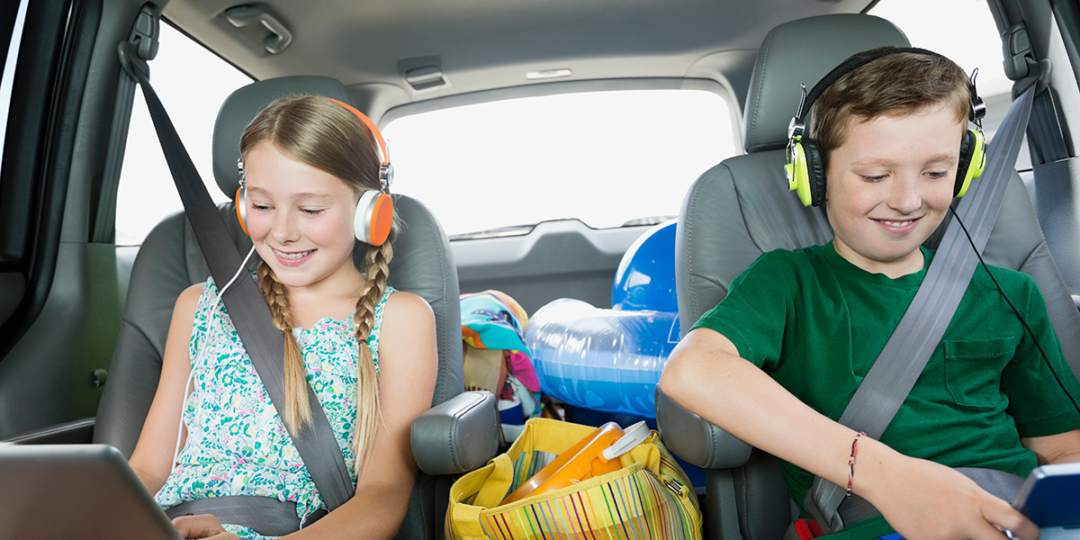 Two middle grade children using devices and headphones in the backseat of a car.