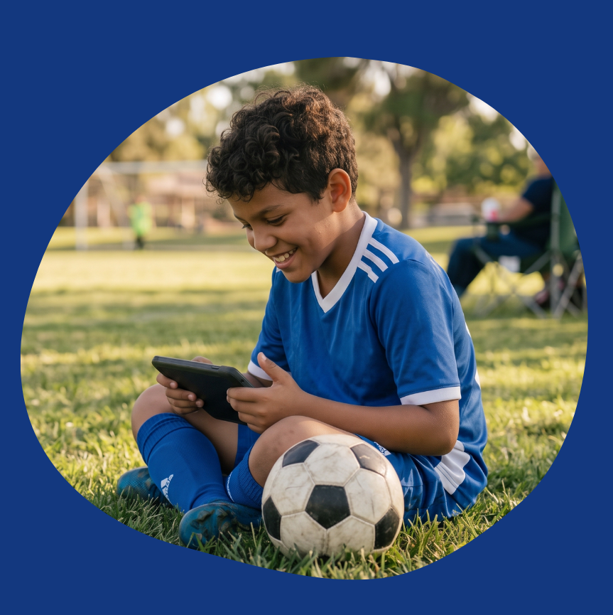 image: a middle-grade aged boy sitting on a soccer field in uniform with a soccer ball next to him and reading on a tablet.